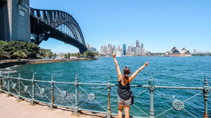 female standing with arms in the air and back to the camera next to the sea with a bridge in the distance